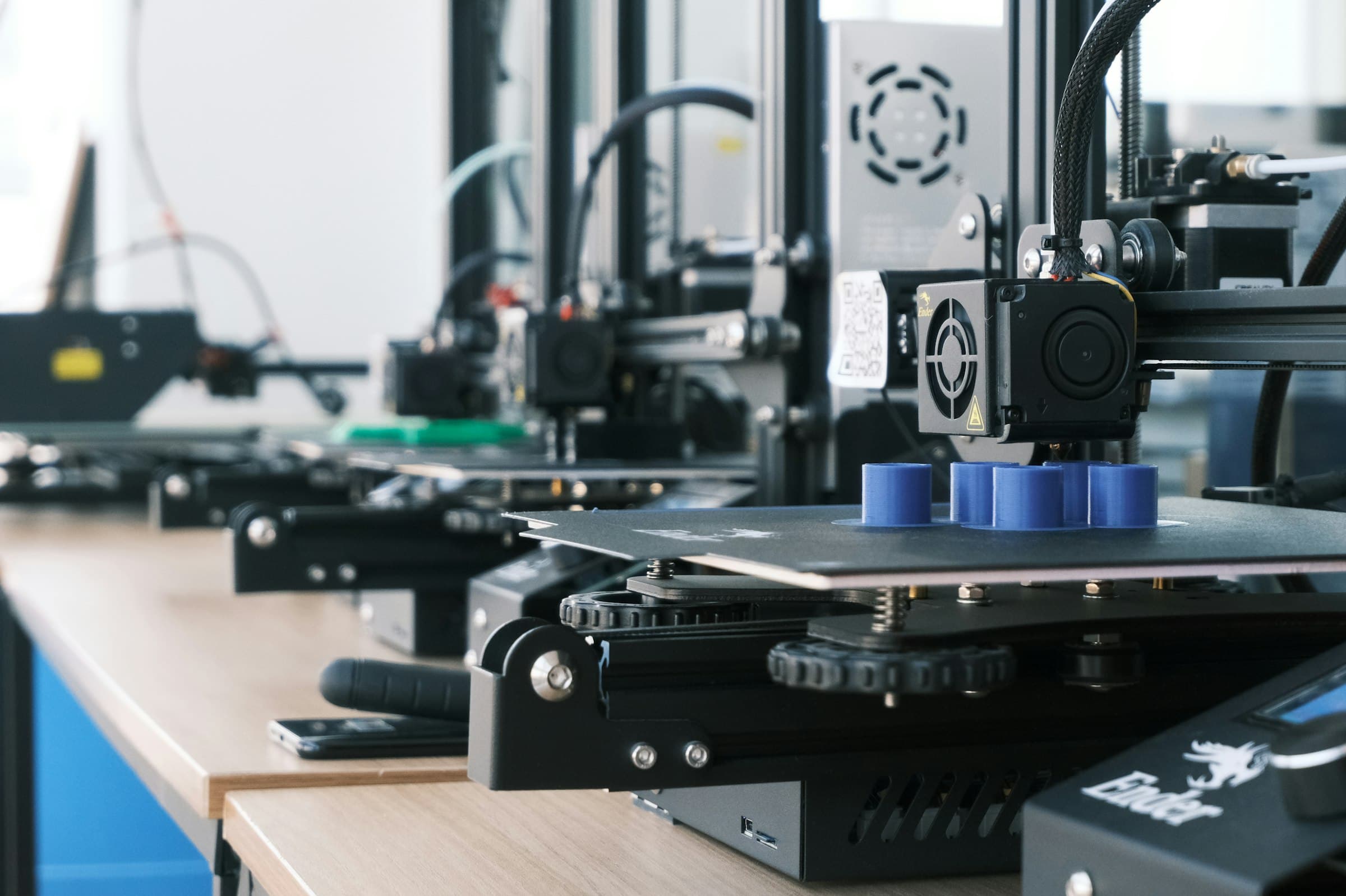 Fileira de impressoras 3D numa bancada, com peças azuis na mesa da primeira